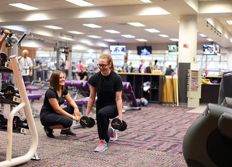 Female personal trainer smiling at a female doing a lunge with free weights in an open area of the fitness center