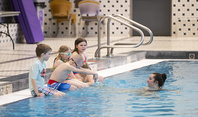 Five children sitting on the side of the pool learning how to dive from an instructor 